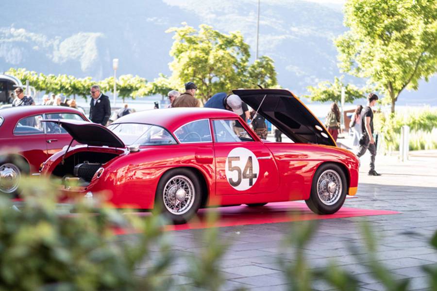 Offene Hauben, gespannte Blicke: Ein roter Sportwagen mit der Startnummer 54 zeigt in der Tessiner Frühlingssonne sein Innerstes – dahinter der Ceresio, davor Fachsimpeleien, Bewunderung und das leise Versprechen von Geschwindigkeit aus einer anderen Zeit.
