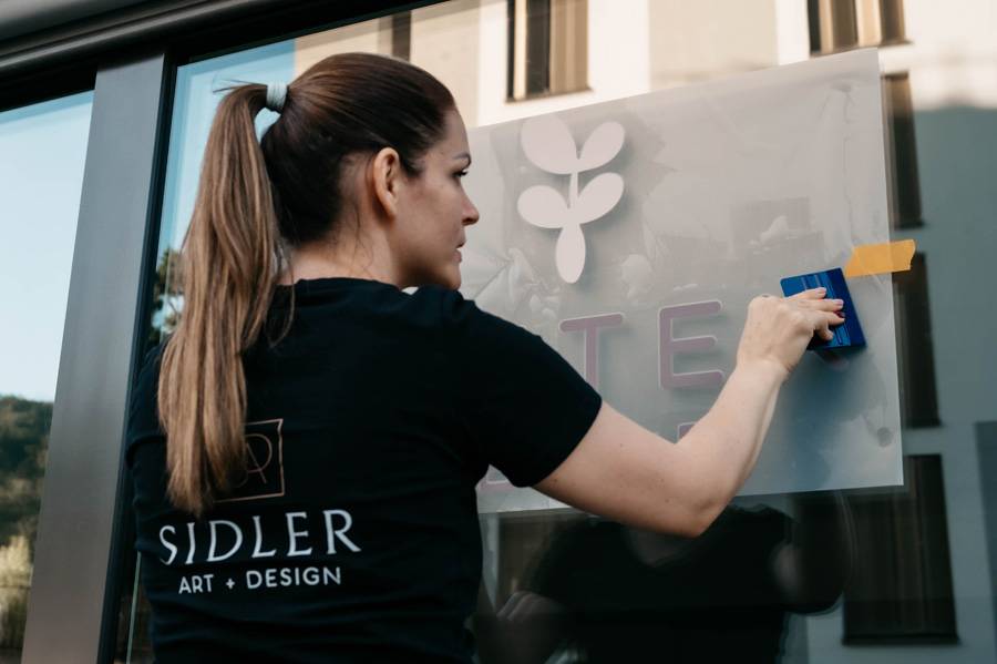 Samira Sidler lors de l’application du film adhésif : geste sûr, regard exercé – le lettrage de vitrine, comme une déclaration visible de la marque et de l’espace.