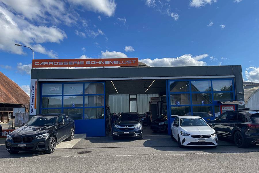 Blau in Bestform: Der Himmel spannt sein schönstes Firmament über den Carrosseriebetrieb, während sich hinter den weit geöffneten Toren der Werkstattalltag zwischen Reparatur, Lack und Kundschaft abspielt.