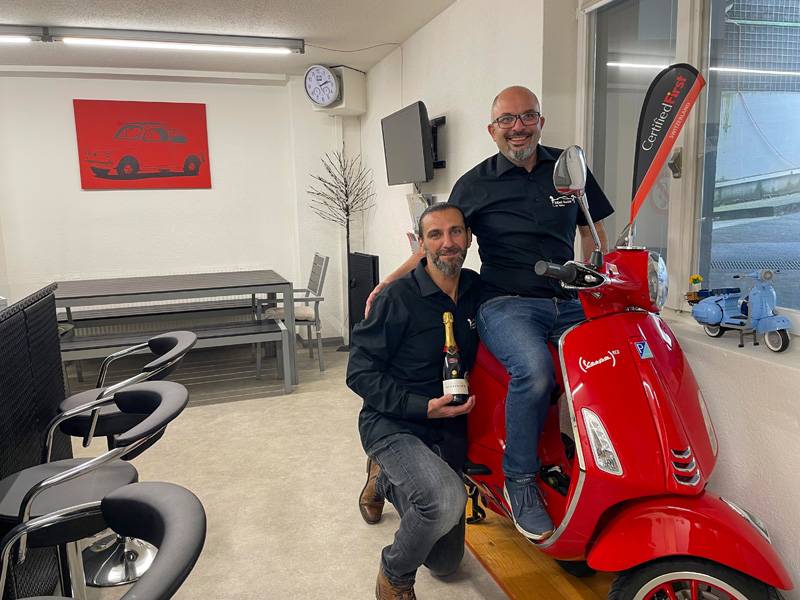 Luca Maggiorelli (à gauche) et Massimo Valvo : associés, beaux-frères et visiblement bien rodés – ici avec une Vespa rouge et un drapeau « Certified First » en charmant décor de fond.