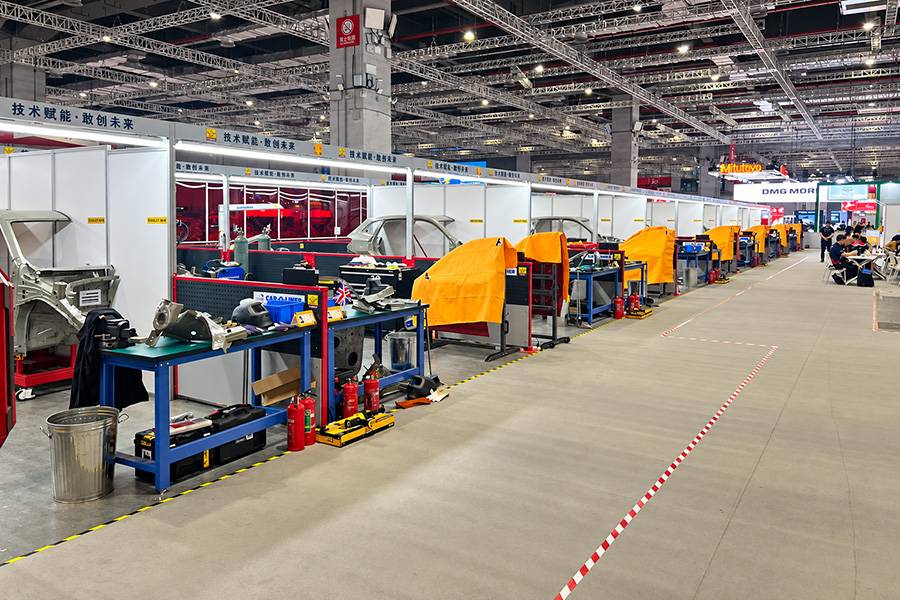 Une longue allée de postes de travail, de chariots à outils et d’éléments de carrosserie, parfaitement alignés comme les stands d’une voie des stands. Chaque concurrent dispose de son propre espace, mais la pression est partout la même : travailler vite, avec précision et sans faute.
